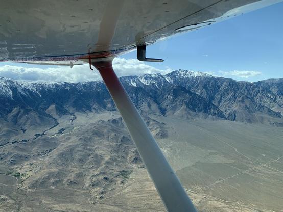 The wing and strut of an airplane in front of  broad desert valley and a snow-capped mountain range. The sky is blue but some cloud hangs above the mountains