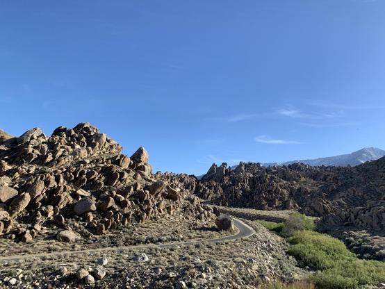 Rocky jagged hills are sunlit under a mostly blue skies. A road curves between them. Taller mountains stand in the distance