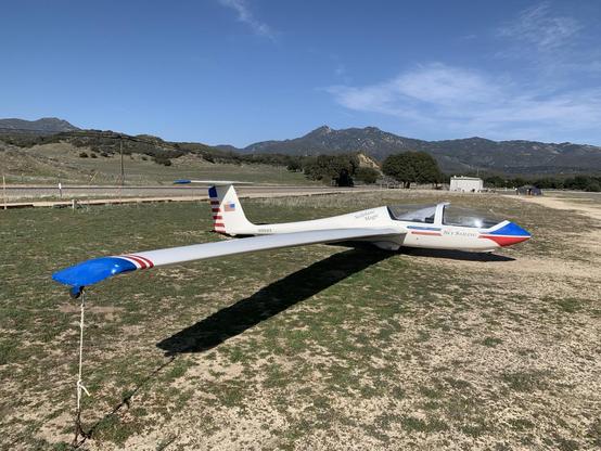A two-seat glider (it’s a Grob 103) stands tied-down in a grassy field on a sunny day