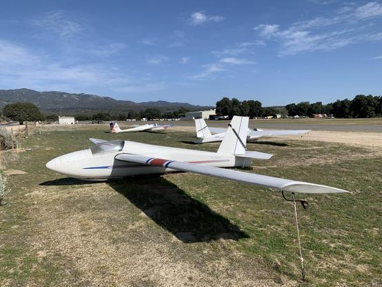 A single-seat glider (it’s a Schweizer 1-26) stands tied-down among other gliders in the grass along a runway.