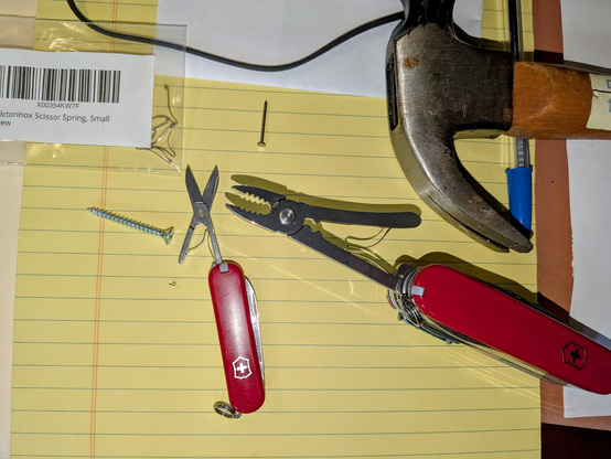 On top of a yellow legal pad is the repaired Victorinox Class SD Swiss Army Knife with its scissors deployed. Next to it is the remainder of the old scissor spring - a stubborn metal curl that had to be hammered out gently using a wood screw as the pushing element. To the right of the repaired Classic SD is a Swiss Champ model Swiss Army Knife with its pliers deployed. Off to the right of the Swiss Champ is a hammer and a blue Bic Cristal ballpoint pen. 

A ziploc bag featuring the replacement Victorinox scissor springs is in the top left corner of the photo. 