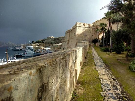White and grey fort tower in the distance, with a similar colour stone wall on the left. A stone path laid on grass leads towards the tower. Trees align the path on the right and sea can be seen on the left with few boats. The sky is clear blue.