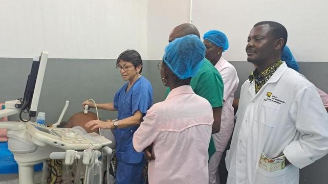Une professionnelle de l’AP-HM en blouse bleue forme un groupe de soignants togolais à l’échographie obstétricale, en présence d’une patiente allongée.