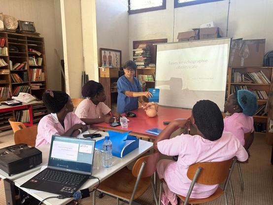 Atelier théorique en échographie obstétricale animé par une soignante AP-HM, face à un groupe de sages-femmes togolaises, avec mannequin pédagogique et support projeté.