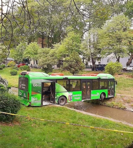 Green "Go Green Hybrid" bus stuck in a shallow stream in a park.