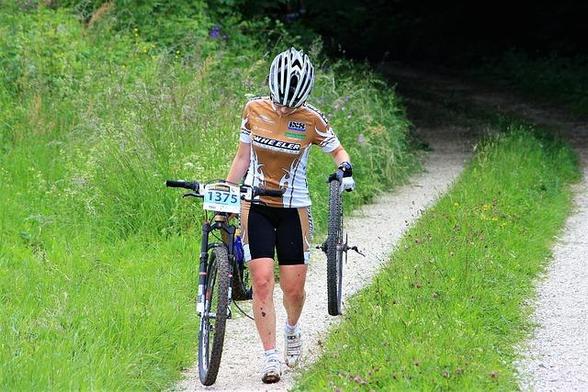 Eine Frau, mit Fahrradtrikot und Fahrradhose sowie Helm auf dem Kopf, läuft auf einem Feldweg umgeben von viel Grün. In der rechten Hand trägt Sie das Hinterrad und in der linken Hand ihr Fahrrad.