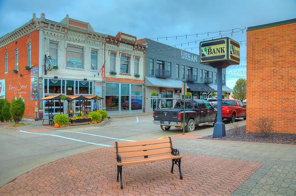 A quaint town square features a bench in the foreground with a view of charming storefronts and parked trucks along the street