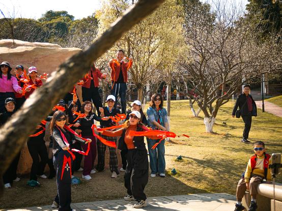 In a sunlit park, a group of people around a large rock are joyfully waving vibrant red ribbons in what seems to be a dance rehearsal. Dressed casually for outdoor fun, with some in hats and sunglasses, they exude energy. In the foreground, a woman in a white cap and dark outfit leads the way with dynamic ribbon-waving. The background, featuring early-spring trees with some leaves budding, has a paved path. A man in dark clothing is seen walking along this path, adding to the scene's everyday charm. Meanwhile, a child in a red vest and sunglasses sits on a stone ledge, perhaps recording the cheerful, communal moment of outdoor merriment.