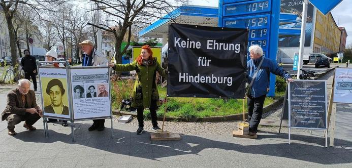 Mehrere Männer und Frauen weisen an einer Kreuzung vor einer Tankstelle auf die Vita Edith Jacobsons hin und präsentieren einen großen Banner mit der Aufschrift "Keine Ehrung für Hindenburg".