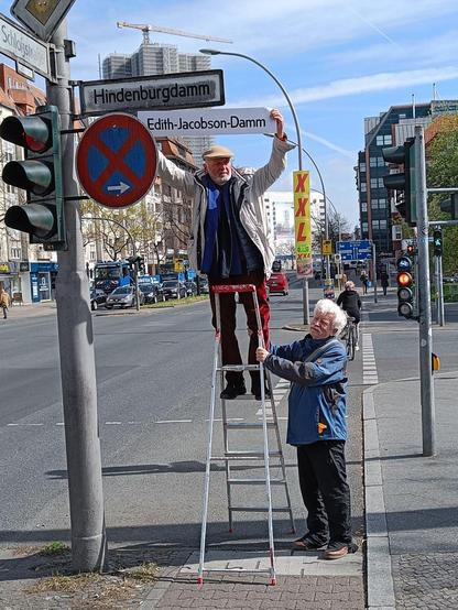 Mit einer Alu-Leiter stehen zwei Menschen neben einem Straßenschild des "Hindenburgdamms", das an einem Ampelmast befestigt ist. Sie halten gleichformatiges Schild mit der Aufschrift "Edith-Jacobson-Damm" darunter.