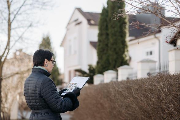 A woman stands in front of a house, holding a piece of paper, with a focused expression on her face