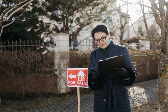 A woman wearing glasses holds a clipboard while examining a real estate sign in an outdoor setting