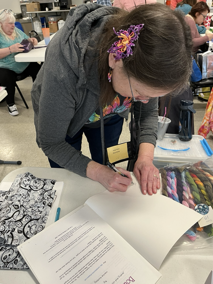That’s me signing one of my tatting pattern books for a customer. Some of my colorful hand dyed thread is also on the table. Some people at the event are partially showing at tables in the background. I’m wearing a brightly colored tatted lace flower in my hair.