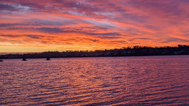 Historical Documentation in the form of a photograph of a rising sun over the ocean bay at the Historic Pioneer Village in Forest River Park, Salem, MA. The sky is filled with vibrant colors of orange, pink, and purple, with the sun rises in the background. The water is calm and reflects the colors of the sky, creating a mirror-like effect. On the right side of the image, there is a small island with trees and houses on it. The island appears to be located on the shore of the water, with a few small islands visible in the distance. The overall mood of the photograph is peaceful and serene as we bear witness to this glorious new day.