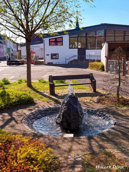 Ein idyllischer Dorfplatz in Laubach-Münster im Frühling: Im Zentrum plätschert ein kleiner Brunnen, umgeben von frischem Grün, einer schattigen Bank und traditionellem Naturstein. Sanftes Licht fällt durch das junge Laub der Bäume und verleiht dem Ort eine ruhige, friedliche Stimmung. Im Vordergrund ist das Dorfgemeinschaftshaus zu sehen, darüber ragt die Spitze des Kirchturms – stille Zeugen gelebten Dorflebens.