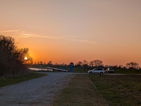 Sonnenuntergang auf einem Schotter-/Rasenplatz am Deich an der Elbe. Weiter hinten stehen zwei PKW, die Sonne ist bald verschwunden. Der klare Himmel ist in Rot- und Orangetönen.