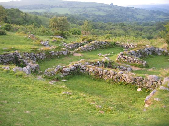 A series of low walls outlining the ruins of several buildings, with farmland in the distance and swathes of green grass in the foreground.