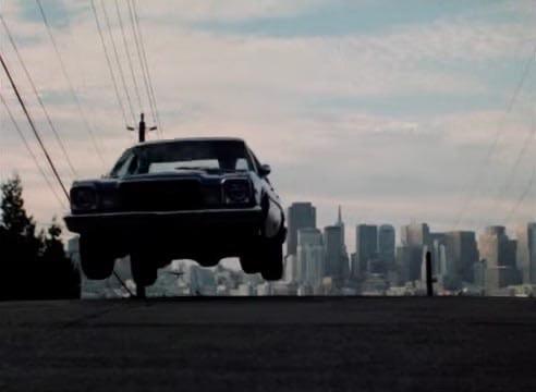 A vintage car is captured mid-air, jumping over a road with a city skyline in the background. Power lines stretch across the image, and the sky is partly cloudy.