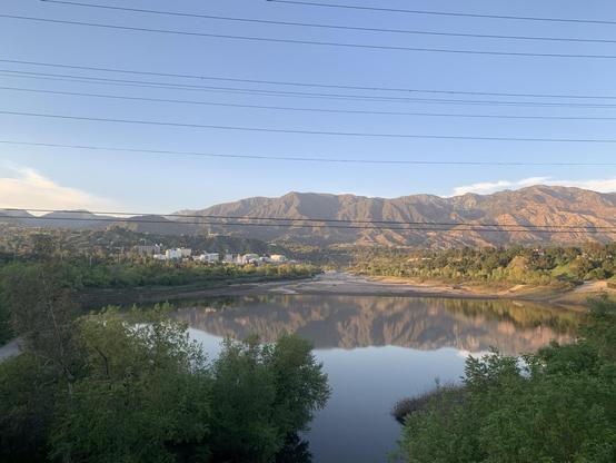 Near sunset, the blue sky and a mountain range are reflected in a calm pony surrounded by trees. At the far end of the pond, a campus of buildings (it’s NASA’s Jet Propulsion Laboratory) stands at the base of the foothills