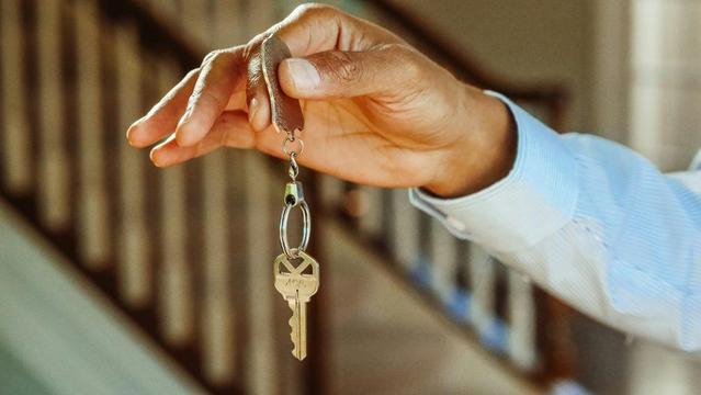 A close-up of a hand gripping a key, representing access to a new home, with a house silhouette nearby