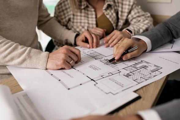 Three individuals examine blueprints spread out on a table, discussing plans and ideas collaboratively