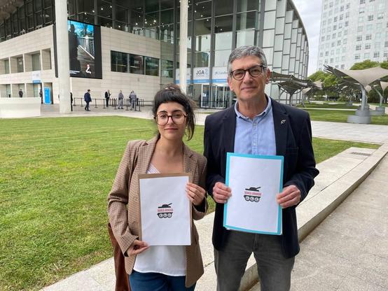 Una mujer y un hombre muestran dos hojas con el logotipo de la campaña banca armada frente al edificio de caixabank, instantes antes de entrar para denunciar sus inversiones en armamento en la junta de accionistas del banco
