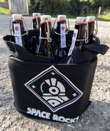 A black cotton tote bag with a stylised astronaut helmet logo inside a diamond frame with the words “Space Rocks” at the bottom. There are six litre bottles of beer with rubber stopper closures in the bag.