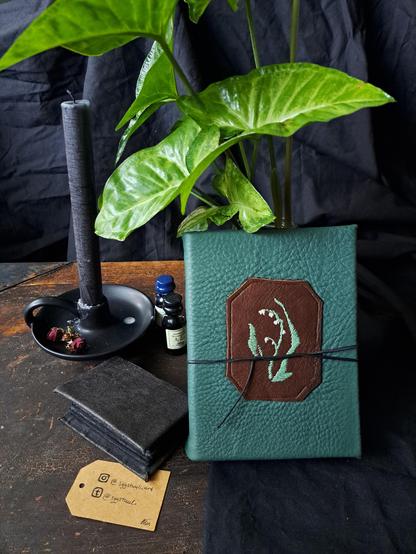 A dark green book cover with a brown leather patch and a flower embroidered in it. The book is leaning on a house plants with big green leaves. There's a small black sketchbook, ink bottles and a black candle on the table next to the book.