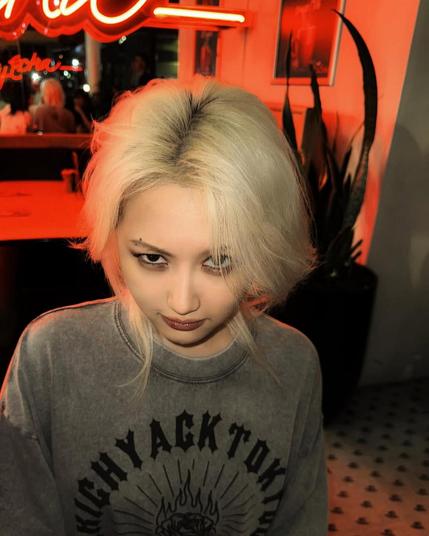 shot of siyeon (dreamcatcher) sitting in a bar, red neon lights, looking into the camera. a fierce and cool look, but still cute. she has wild blonde hair and is wearing a grey shirt.
