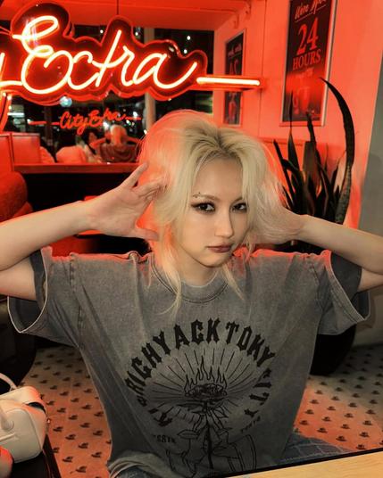 shot of siyeon (dreamcatcher) sitting in a bar, red neon lights, posing and looking into the camera. a fierce and cool look, but still cute. she has wild blonde hair and is wearing a grey shirt.