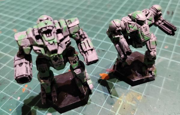 Two OmniMech minis, a Kingfisher and a Nova Cat, painted grey with green stripes for Clan Jade Falcon's Gamma Galaxy are placed on a well-worn cutting mat. All details and bare metals are yet untouched.