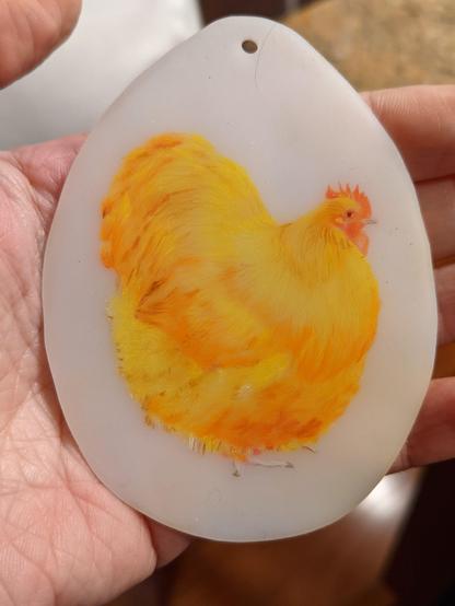 Photo of a left hand holding an egg-shaped keychain made of shrinky dink plastic. In the middle is a hand drawn fluffy chicken drawn with colored pencil.