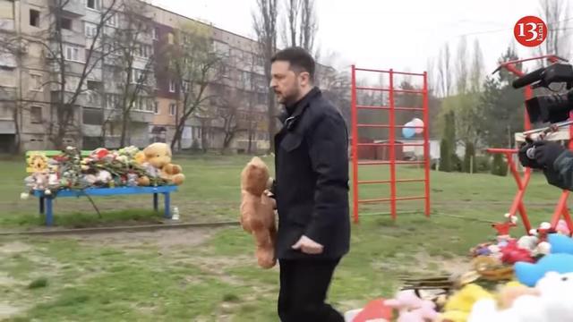 President Zelenskyy is visiting the playground carrying a brown teddy bear. In the background people have placed teddy bears and flowers on a bench which has become a site of mourning.