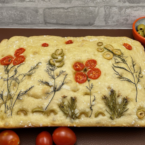Homemade focaccia with tomatoes, olives and herbs arranged to form flowers. 
