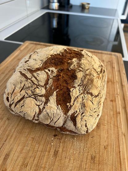 A loaf of freshly baked bread resting on a wooden cutting board. Nice brown colored crust covered in flower. A clear, deep crack going through the middle. 