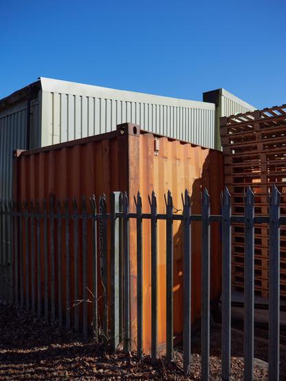 Metal fence with orange containers behind it. Behind those a grey warehouse roof and a stack of palettes to the side.