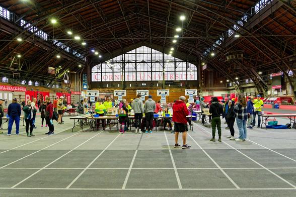 Here is a big hall with a triangle topped with that says CORNELL UNIVERSITY in Red Letters with banners that say AIR FORCE and ARMY on the side at the back of a huge open indoor tracks with dramatic arches, glowing lights, a row of windows stretching towards the viewer, catewalks and more signs for Cornell athletics.

Looking tiny in comparsion is a reception area made of temporary tables with capacity to handle five lines of alphabetically sorted runners which are now empty while various people mill around.