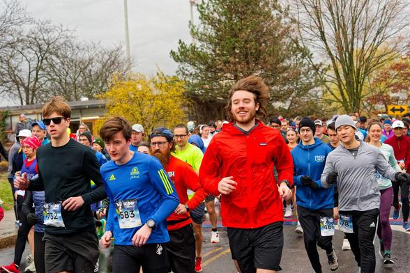 A crowd of a few hundred runners breaks from the starting line,  most notably a man in a red jacket with brown windswept hair.  Behind find a random but pleasing mixture of trees and shrubs.
