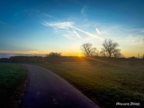 Ein schmaler Weg schlängelt sich durch eine weite Landschaft im Abendlicht. Die Sonne steht tief und taucht Himmel und Felder in warmes Gold. Am Horizont zeichnen sich sanft Hügel und Bäume ab. Die Szene wirkt ruhig, offen und einladend – wie der Beginn einer stillen Reise ins Ungewisse.