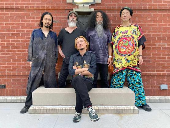 The photo features the five current members of Acid Mothers Temple & The Melting Paraiso U.F.O. posing for a photo, with Jyonson Tsu sitting in front of the rest.  