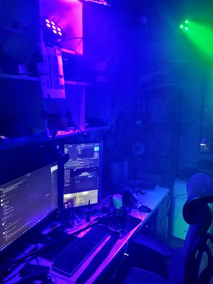 A desk with 2 monitors plunged in smoke with a violet party light positioned above the desk projecting onto a reflector which bounces the light downwards, and a another green spotlight shining at a wall behind the chair. Vertical shot.