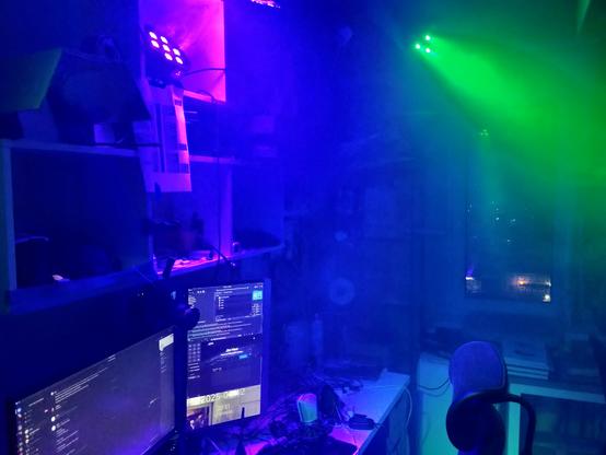 A desk with 2 monitors plunged in smoke with a violet party light positioned above the desk projecting onto a reflector which bounces the light downwards, and a another green spotlight shining at a wall behind the chair. Horizontal shot.