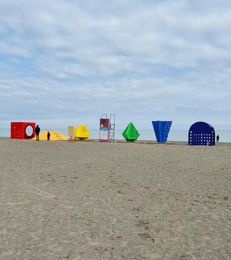 A beach scene featuring large colorful geometric climbing structures in various shapes, including a circle, triangle, square, and more. Two people are seen near the structures, with the lake in the background under a cloudy sky.