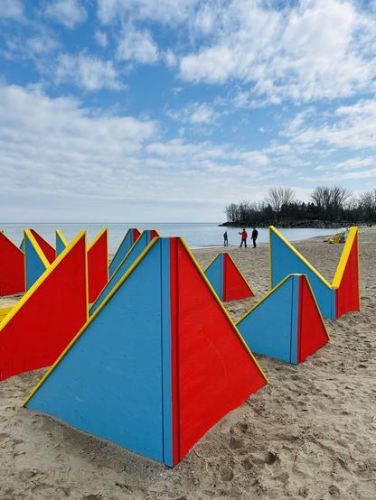 Colorful geometric structures in red, blue, and yellow are arranged on a sandy beach. In the background, people walk along the shore with a view of the water and a tree-lined island under a partly cloudy sky.
