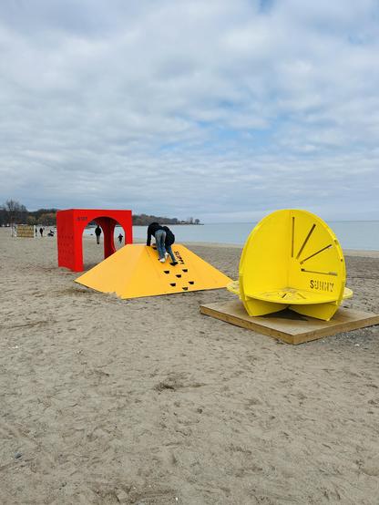 The image shows a sandy beach with three large, colorful geometric structures. A person is climbing on a yellow pyramid-shaped structure, while a red arch and a yellow circular structure are nearby. The beach is relatively quiet, with a few people in the background