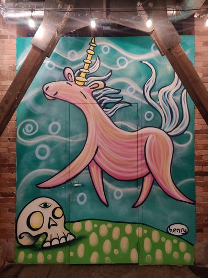 An interior photo of a graffitied wall, featuring a happy pink unicorn framed by two gravy iron supportive structures. Toward the bottom of the piece, there's a skull with 3 eyes.