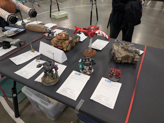 Many game miniatures of vehicles displayed for competition with a sign showing it's the "Space & Sci-Fi D3) Gaming Vehicles" category.  Mostly Warhammer 40k tanks and what looks to be large power armour/small mechs. but there's are also some two battletech minis for an H-7 Warrior VTOL and a Maxim hovercraft both in First Taurian Lancers colours.  A sign reads "Gaming Vehicles"