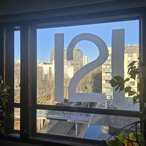 View from a window in Eastern Pasila, Helsinki. Outside is a blue sky, several concrete buildings, and tram tracks. Inside are plants. The number 121 cut out of paper is taped to the window.

The image is mirrored, so that the number can be read from the inside rather than the outside.