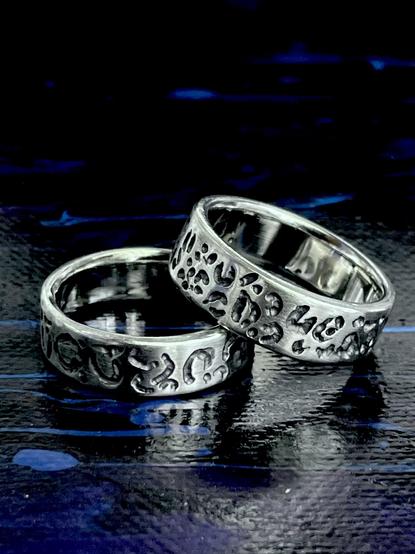 Two handmade silver rings are shown, one featuring a smooth surface and the other with a textured, patterned design. The rings rest on a dark, reflective surface that enhances their shine.