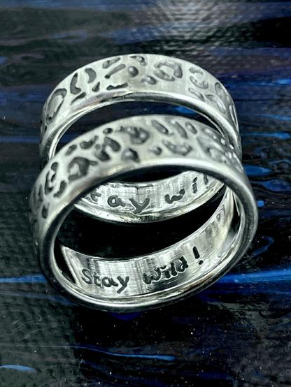 Two silver rings are stacked on top of each other. The rings feature a textured, animal print design and show the engraved phrase "Stay Wild!" on its inner surface. The background is a dark, glossy surface with hints of blue.